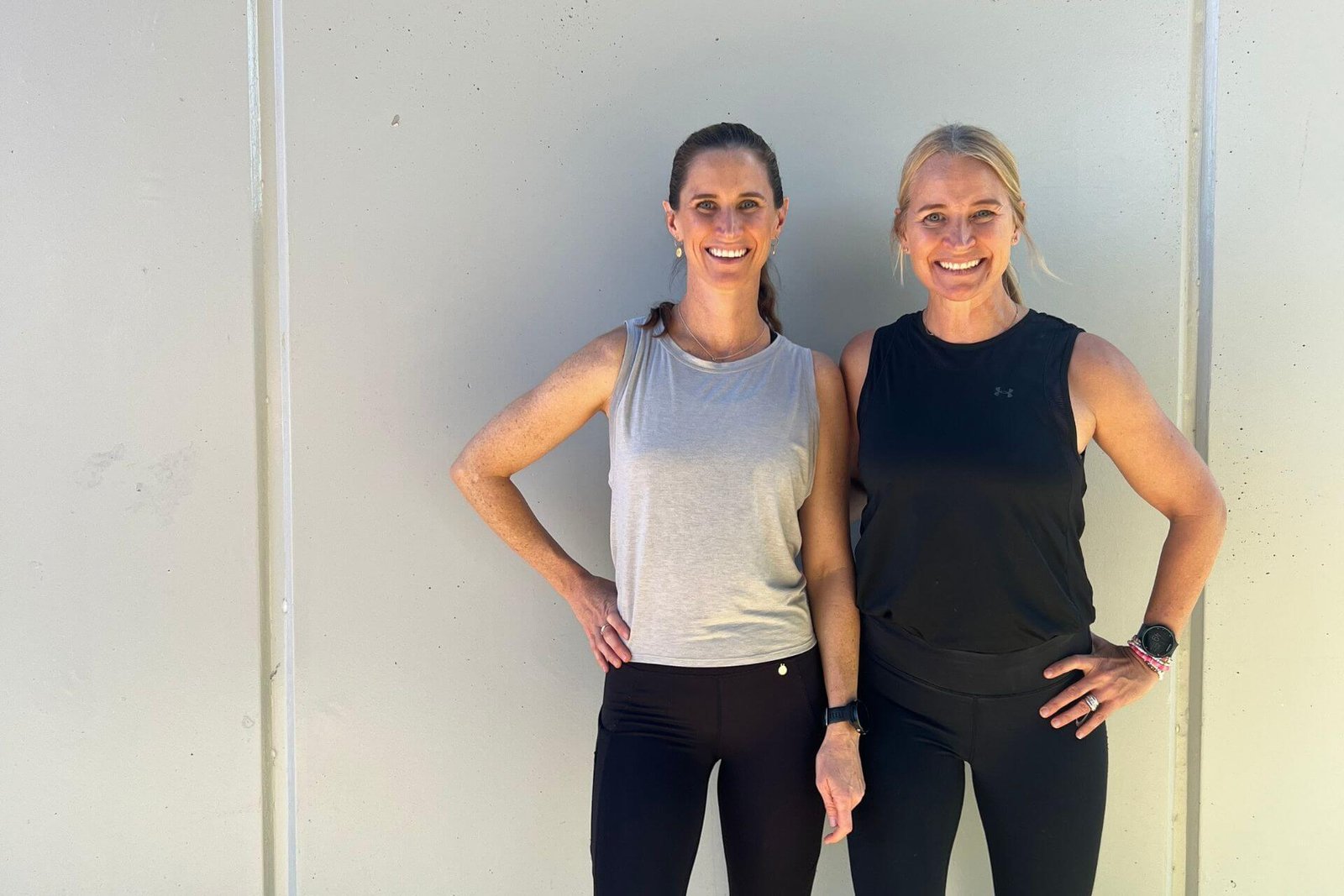 Anna and Em standing in front of a lined grey wall smiling