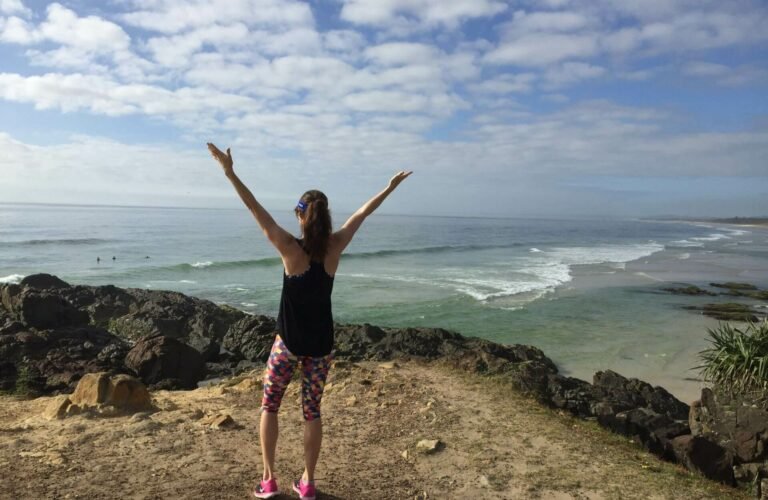 Em raising arms to the sky from a beachside clifftop